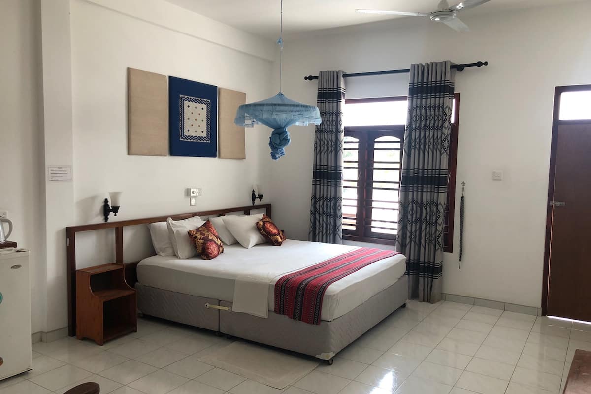 Une chambre à coucher comprenant un lit, un ventilateur de plafond, une fenêtre avec des rideaux et un luminaire fixé au mur.