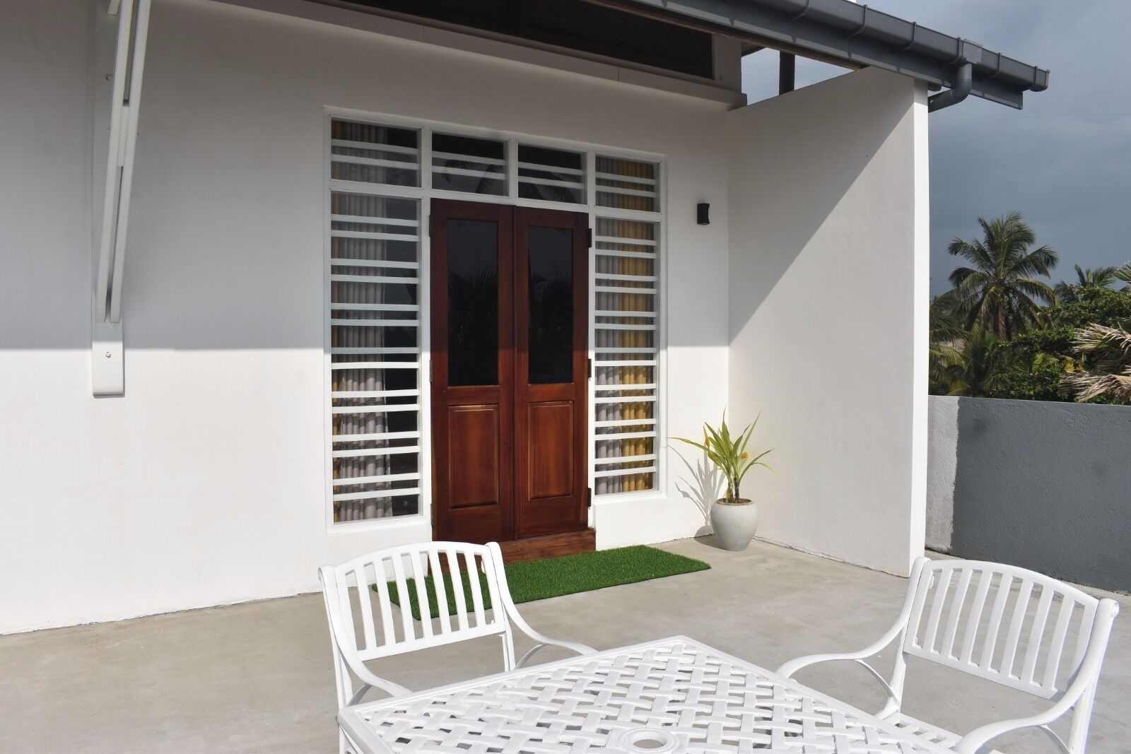 A patio with two white metal chairs and a table, a wooden door with glass panels, and a potted plant.