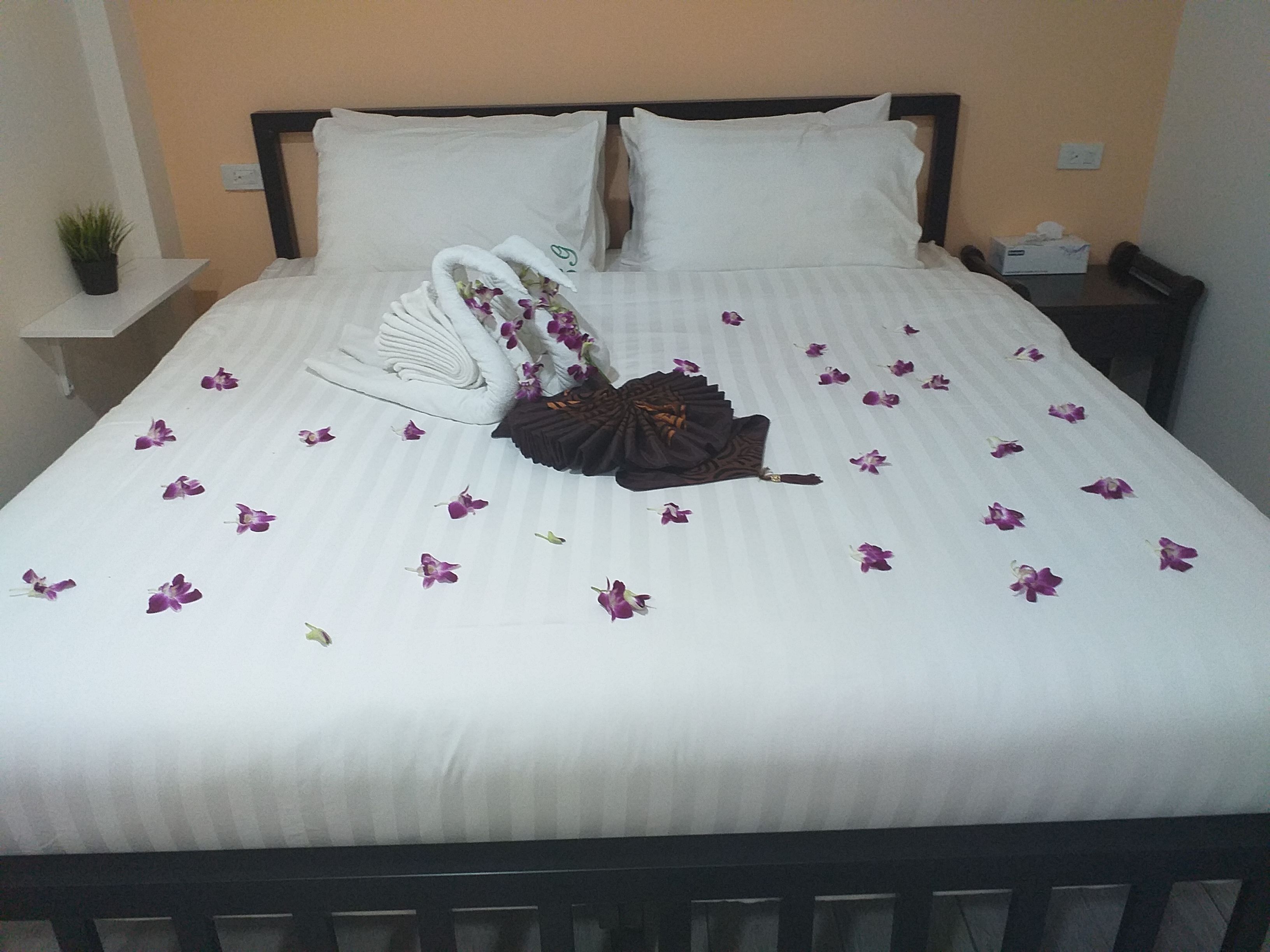 A neatly made bed with white linens, a decorative swan pillow, and scattered purple flowers.