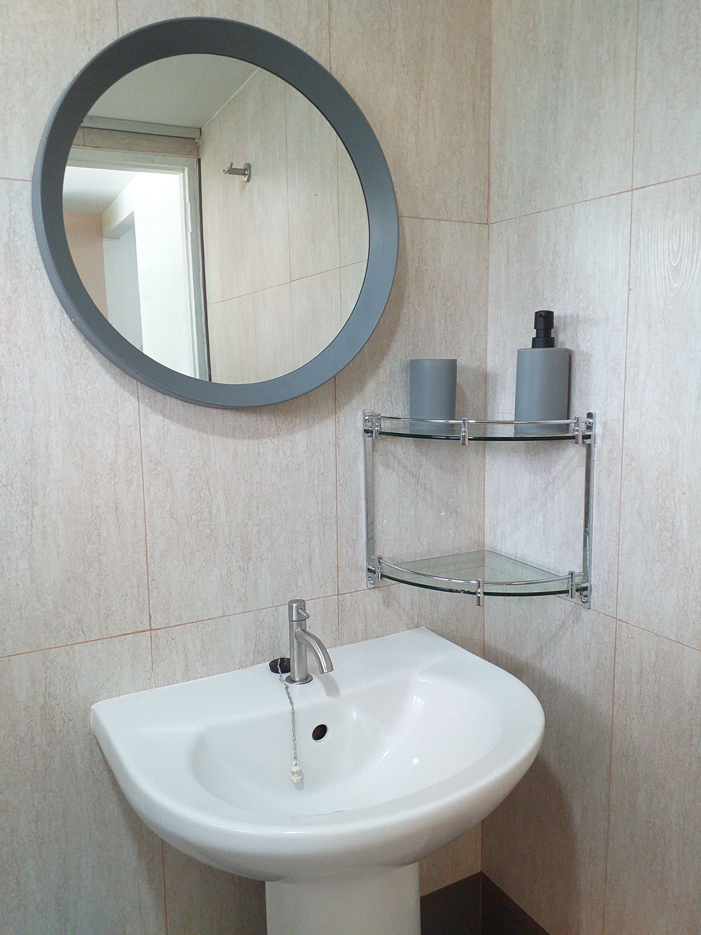Une salle de bain avec un lavabo blanc, un miroir rond et une étagère en verre où sont rangés les articles de toilette.