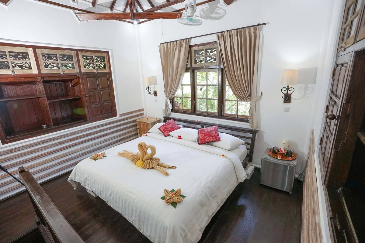 A neatly arranged bedroom with a bed, wooden cabinets, a window with curtains, and a small table with a lamp.