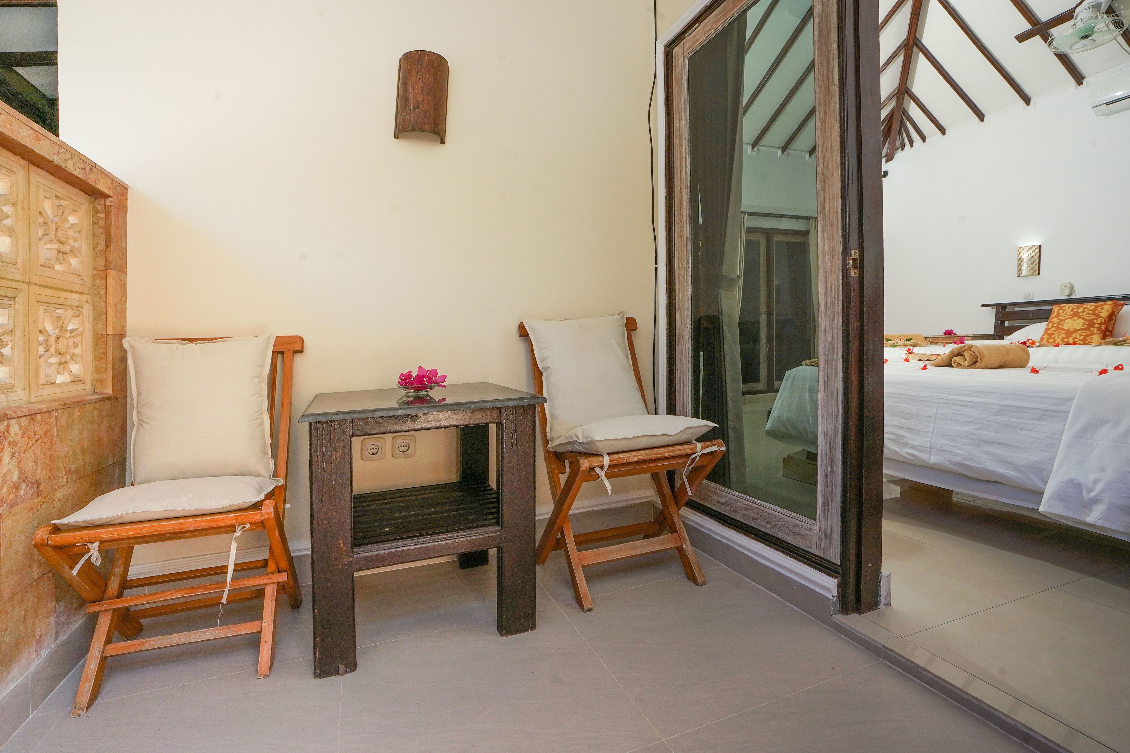 A balcony with two wooden chairs, a small table with a vase of flowers, and a bed with a white bedspread and pillows.