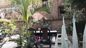 Un patio con mesa y sillas, rodeado de plantas en macetas y un muro de ladrillo.