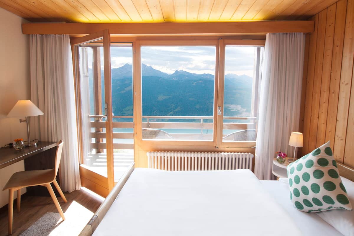 A hotel room with a bed, a desk, a chair, a window with a mountain view, and a balcony.