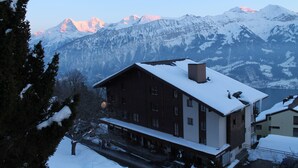 Un edificio cubierto de nieve con un balcón, rodeado de árboles nevados y montañas al fondo.