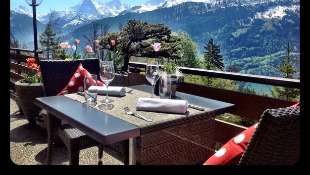 Mesa para dos con vista a la montaña, sillas de mimbre y un florero con flores.