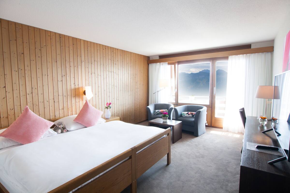 A hotel room with a wooden paneled wall, a bed with pink pillows, a sofa, a desk with a lamp, and a view of mountains through the window.