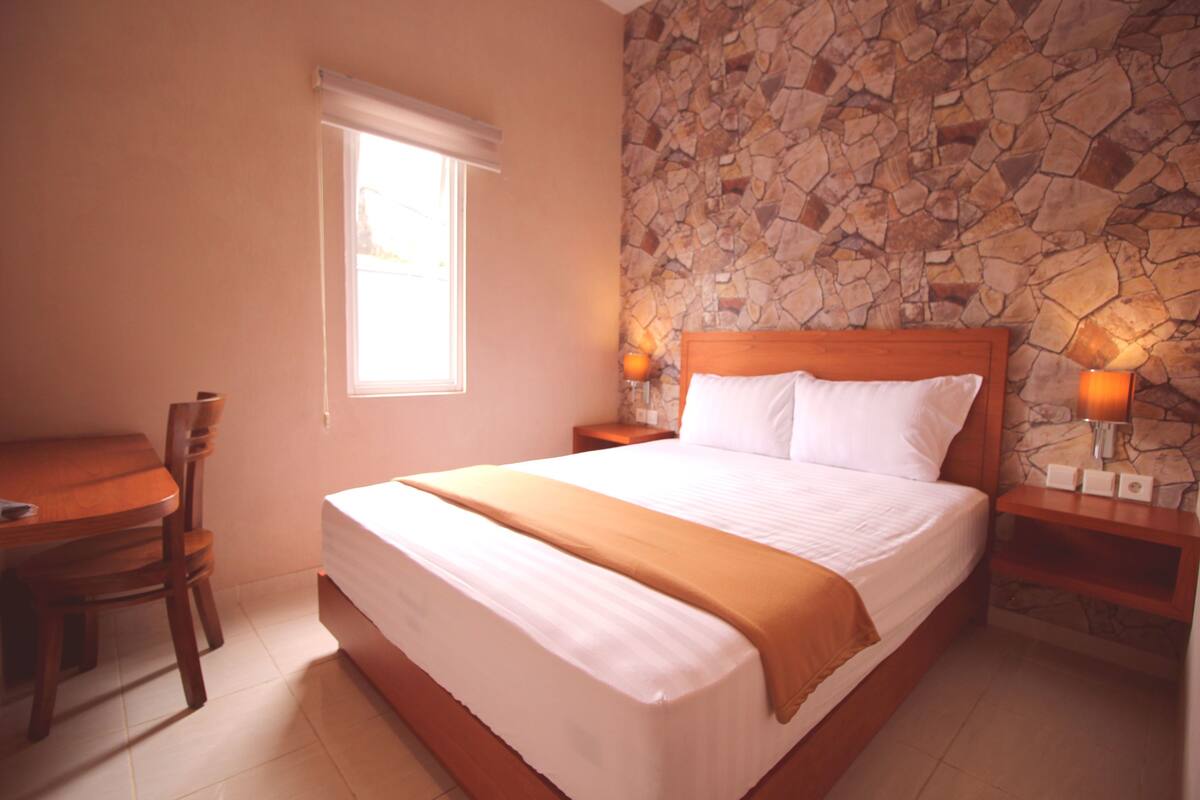 A bedroom with a stone wall, a wooden bed, a bedside table with a lamp, and a window with blinds.