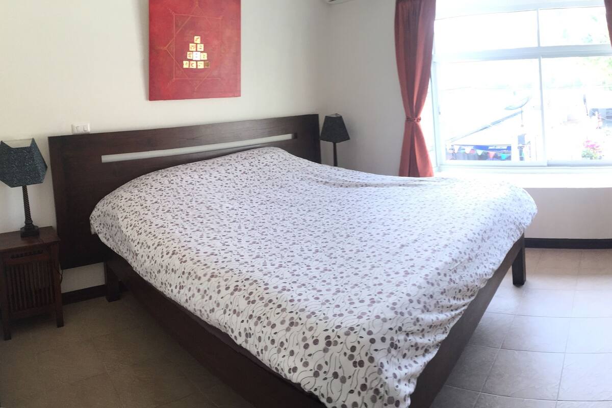 Una cama bien hecha con una colcha estampada con motivos florales en una habitación con un ventanal grande, un cabecero de madera y una cortina roja.