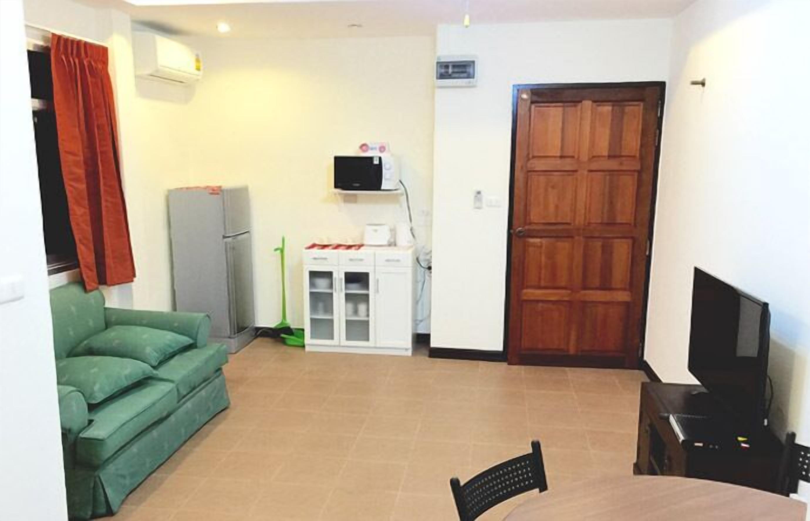 Uma pequena sala de estar com um sofá verde, uma porta de madeira, uma televisão e uma cozinha com geladeira e micro-ondas.