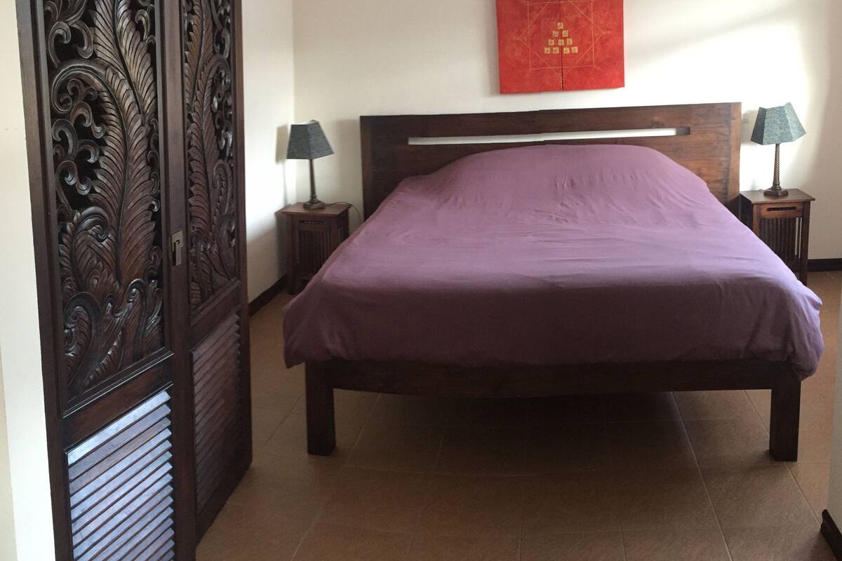 Un dormitorio con una cama, dos mesitas de noche, un aire acondicionado de pared y un panel decorativo a la izquierda.