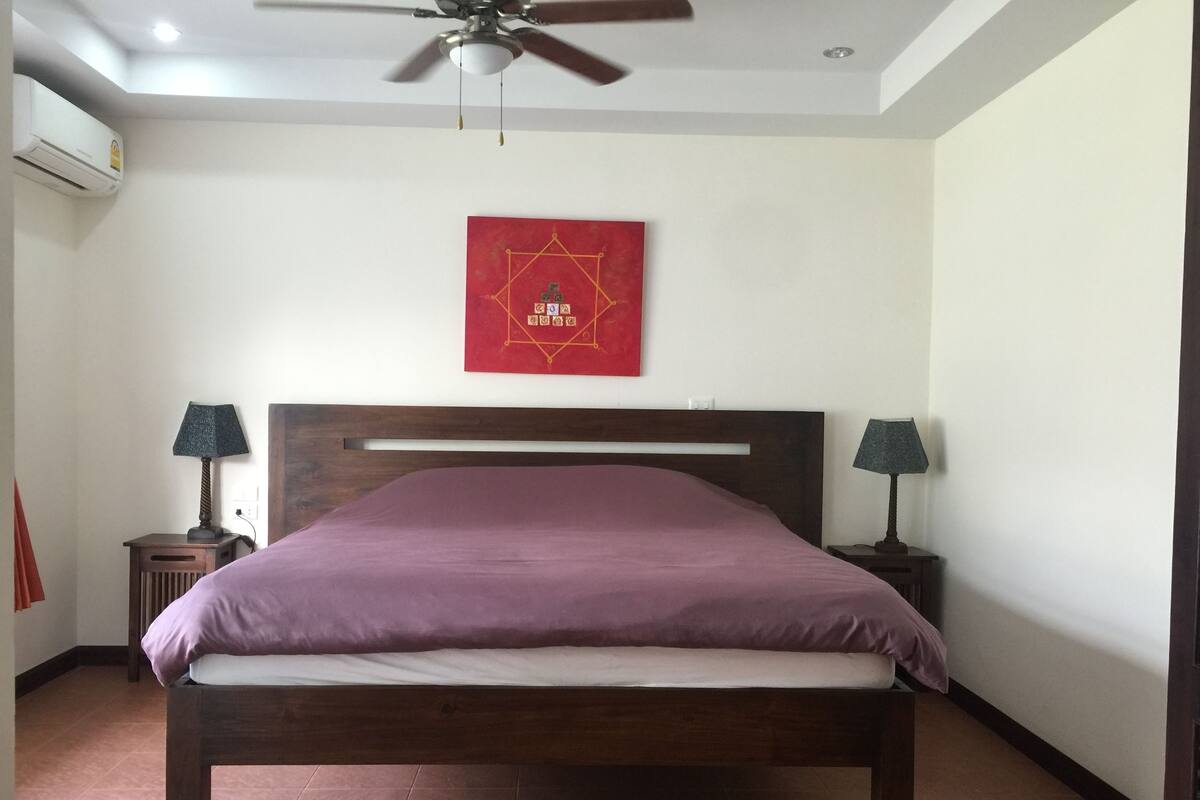 Un dormitorio con una cama de madera, dos mesitas de noche con lámparas, un ventilador de techo y un adorno rojo en la pared.