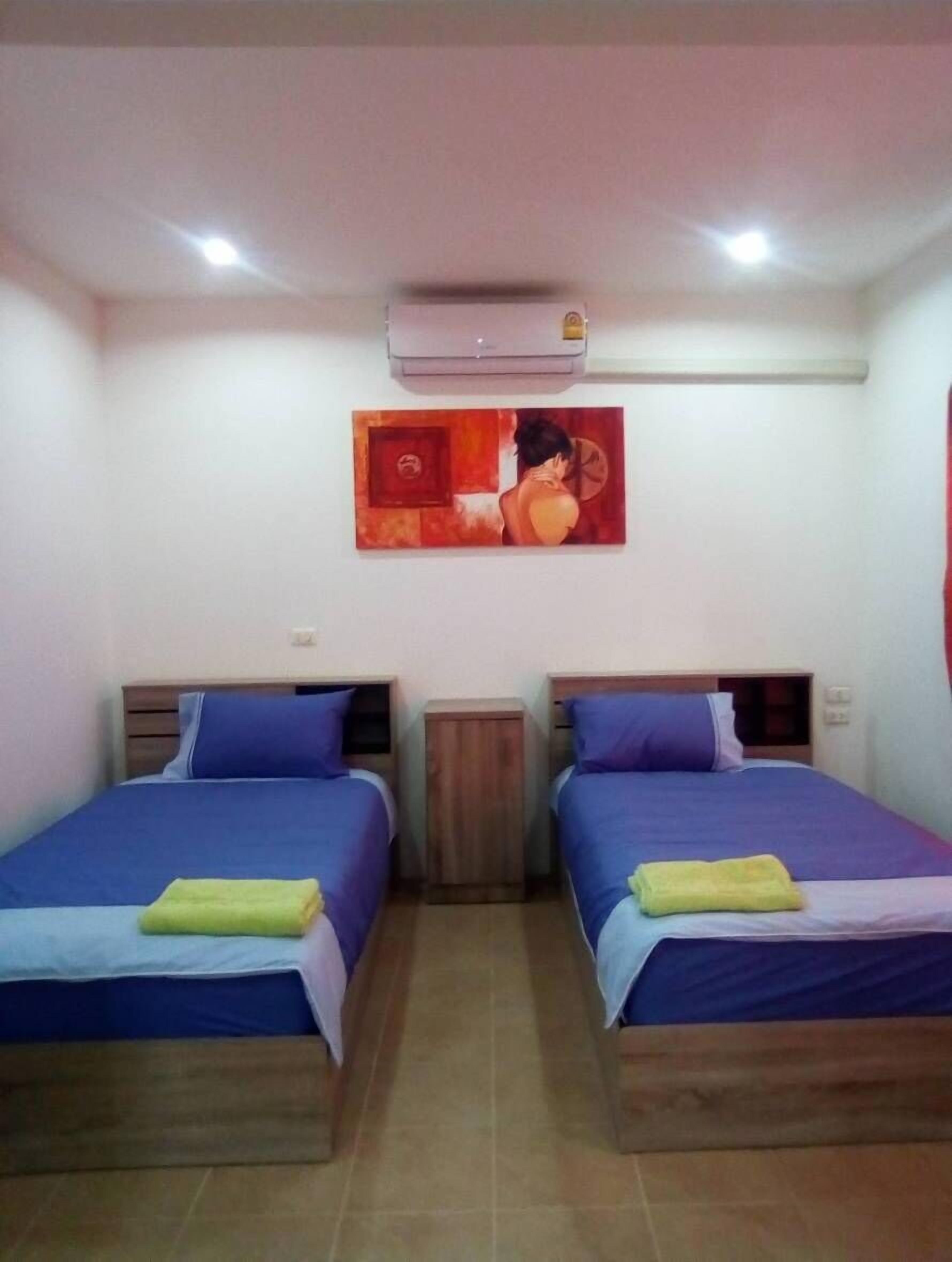 A room with two beds, each with blue bedding and a pillow, a small wooden nightstand, a wall-mounted air conditioner, and a painting above the beds.