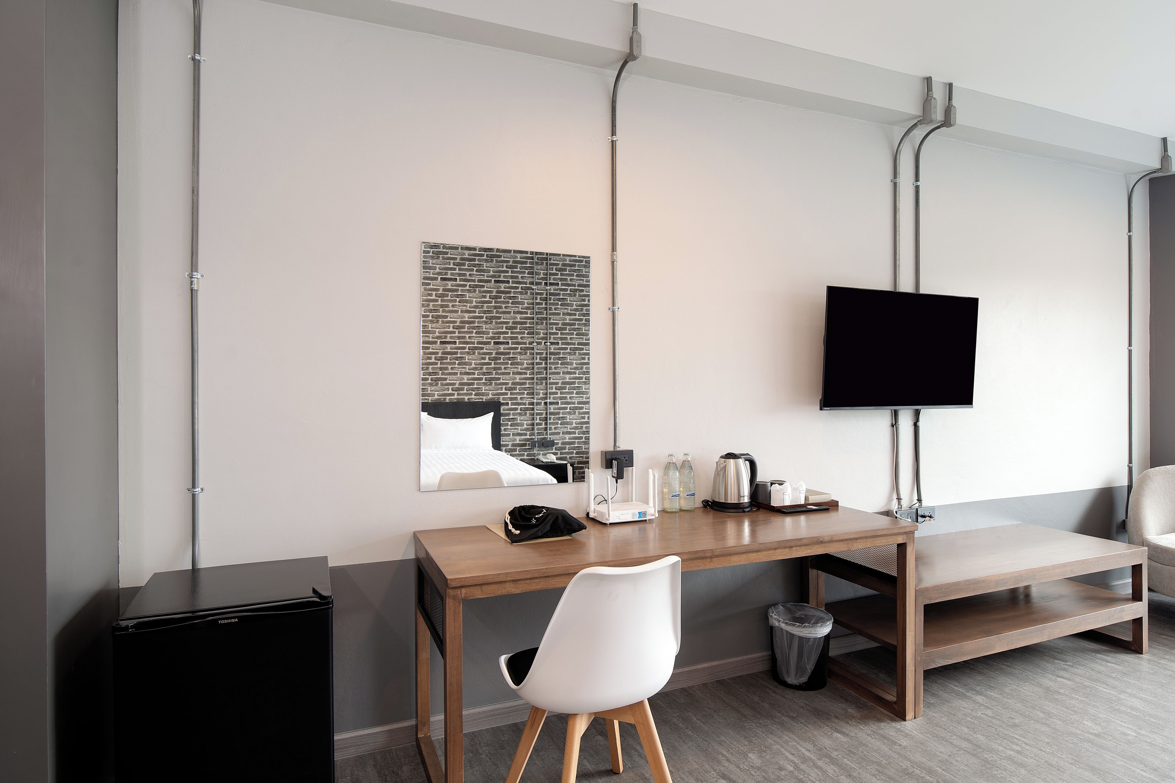 A modern hotel room with a wooden desk, a white chair, a black trash bin, a flat-screen TV mounted on the wall, and a mirror reflecting the room.