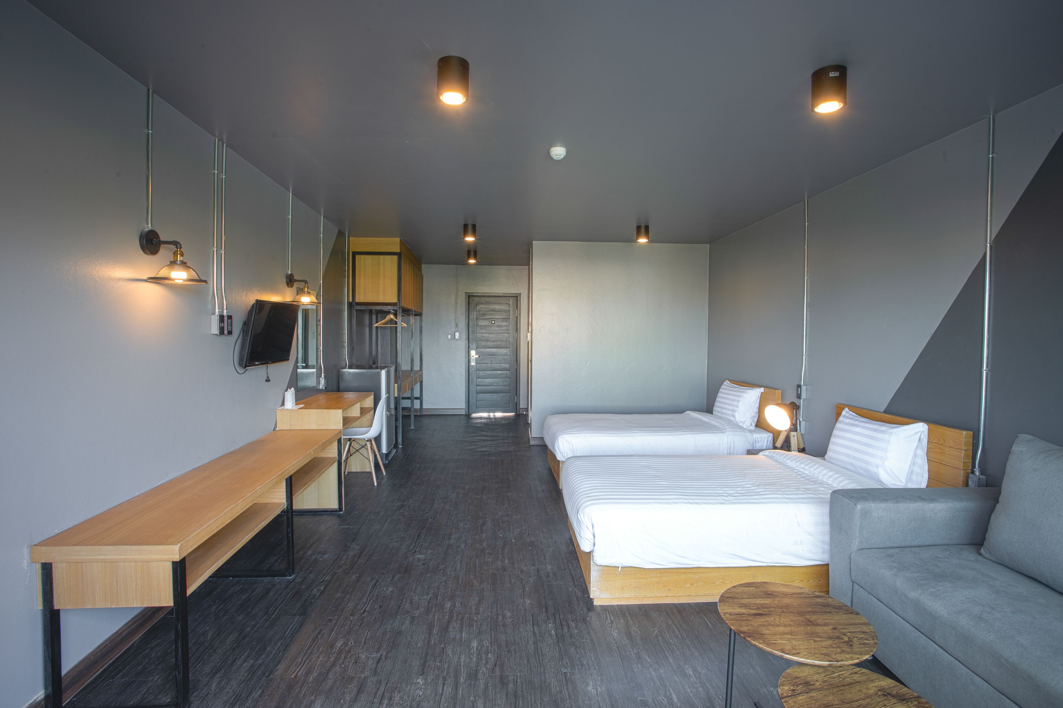 A modern hotel room with a long wooden desk, two beds, a grey sofa, and a small round table.