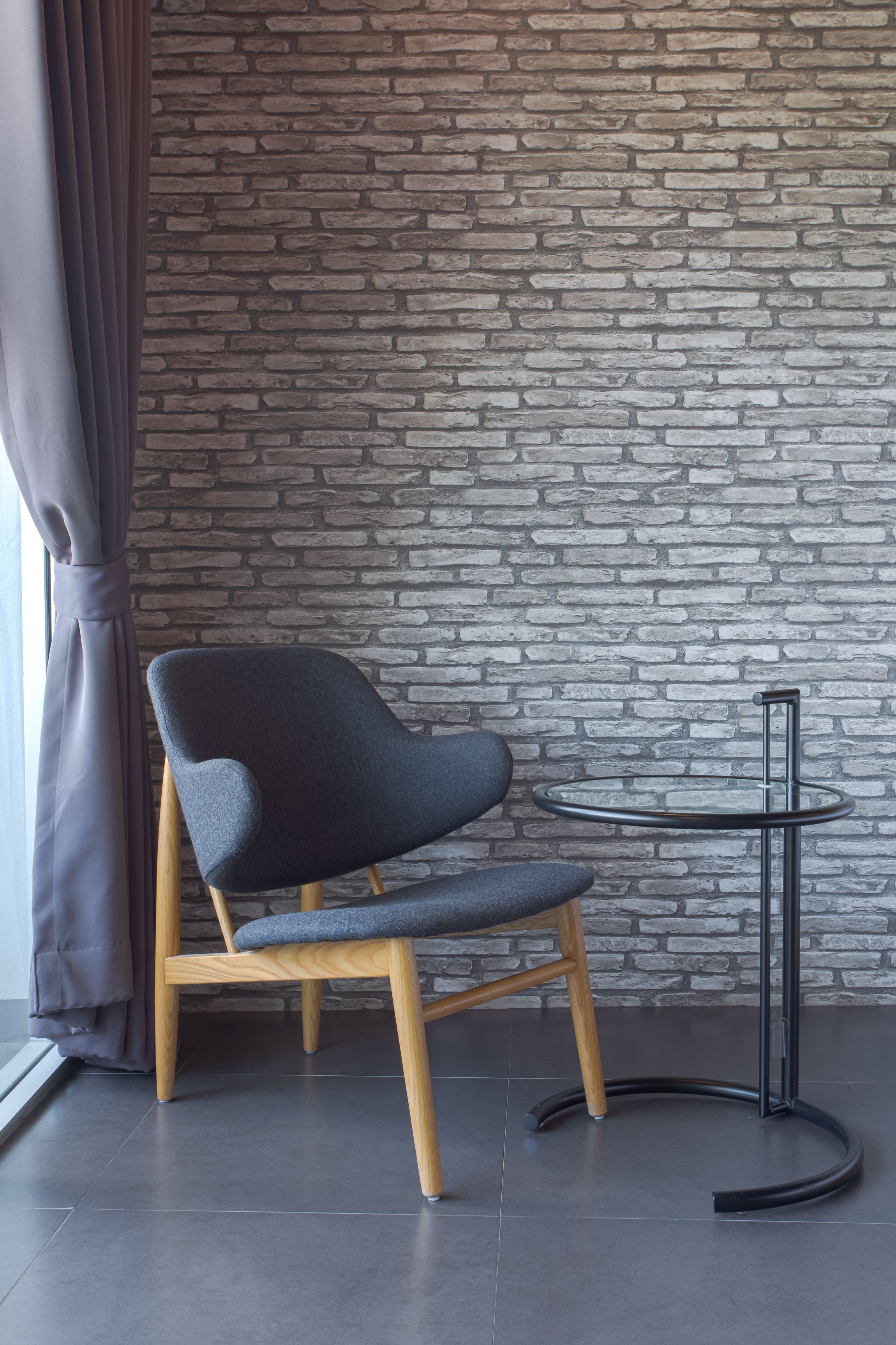 A modern chair with wooden legs and a grey cushion, positioned next to a small round table with a black metal frame, against a brick wall.