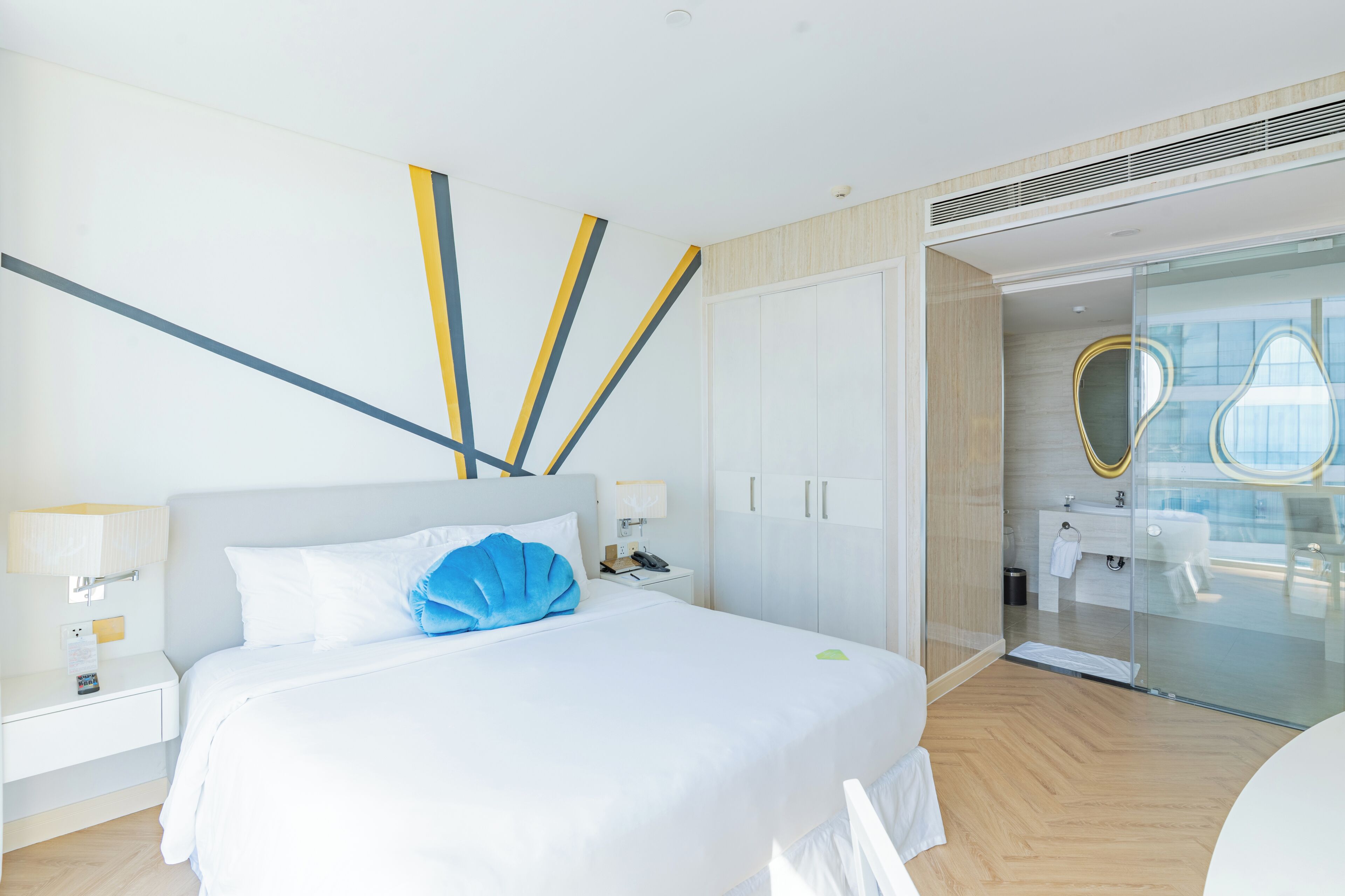A modern hotel room with a large bed, a bedside table, a nightstand, a glass shower enclosure, and a bathroom visible through the glass door.