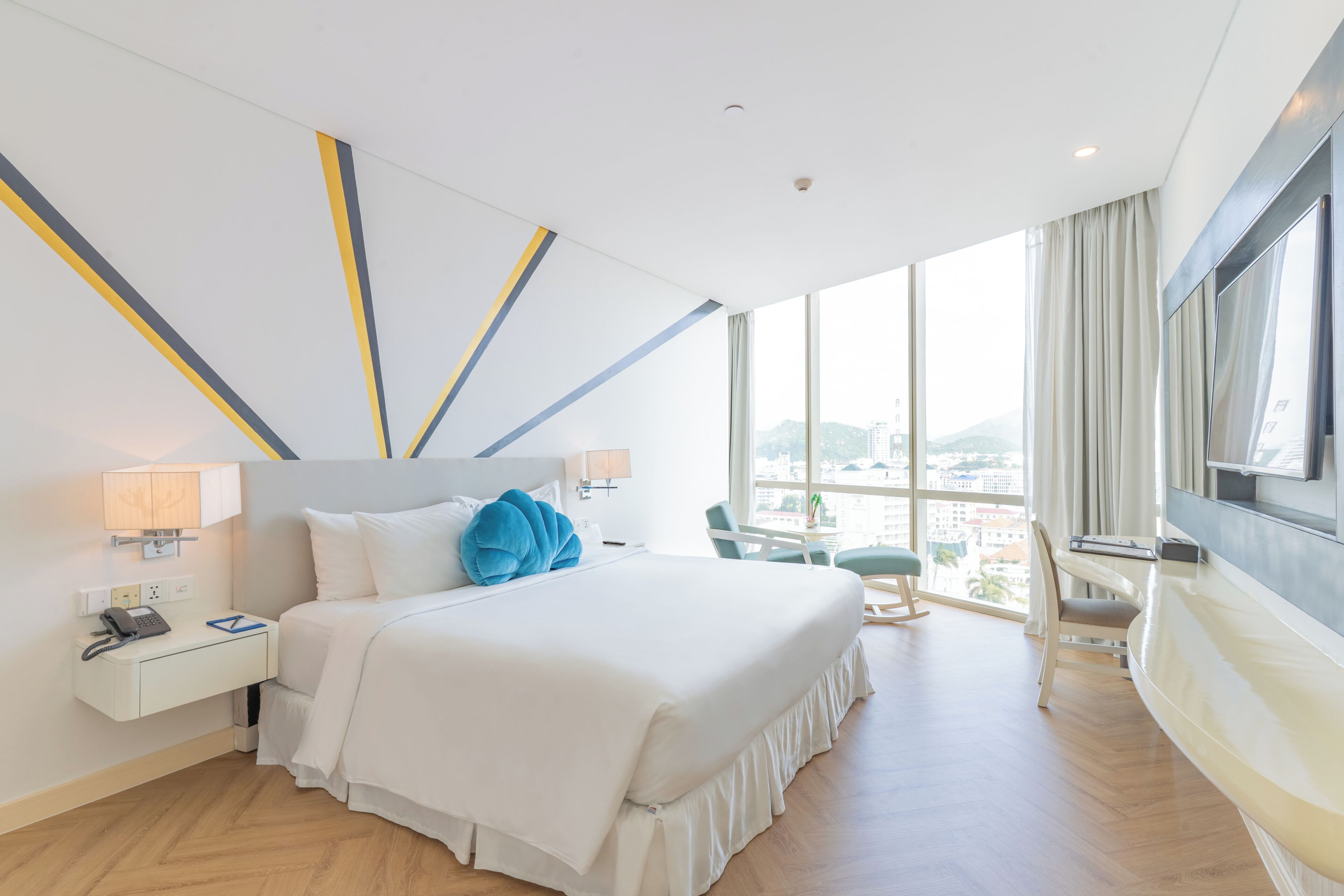 A modern hotel room with a large bed, a desk with a lamp, a chair, and a large window offering a city view.