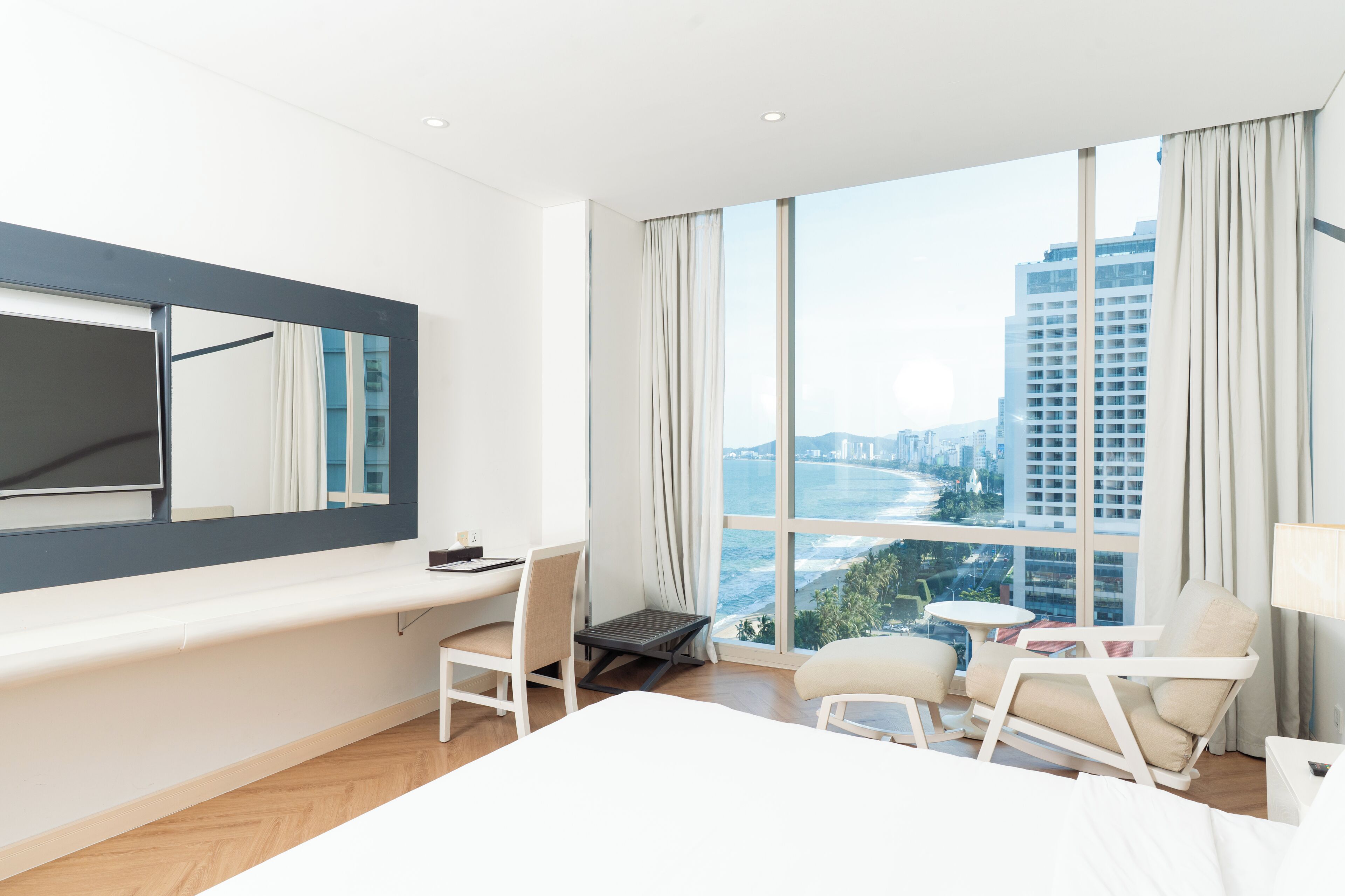 A hotel room with a large window, a bed, a desk, a chair, and a small table.