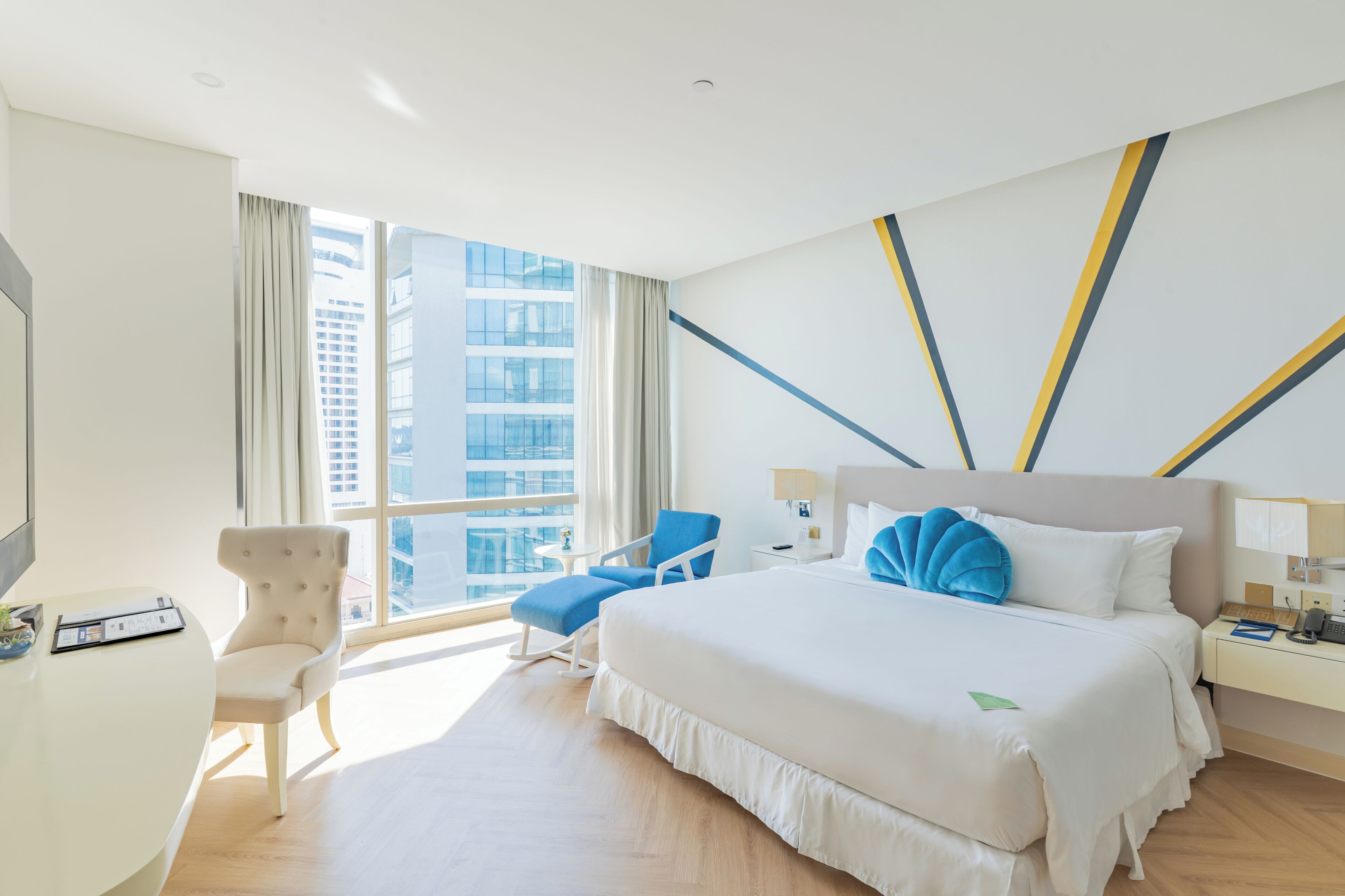 Habitación de hotel con una cama grande, un escritorio, una silla, una mesita de noche y vistas a un paisaje urbano a través de amplias ventanas.