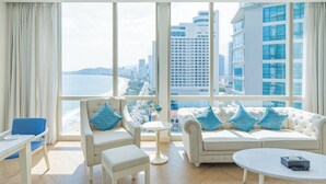 A modern hotel room with a large window, a white sofa, a blue armchair, a small round table, and a white coffee table. - Diamond Bay Hotel (Nha Trang)