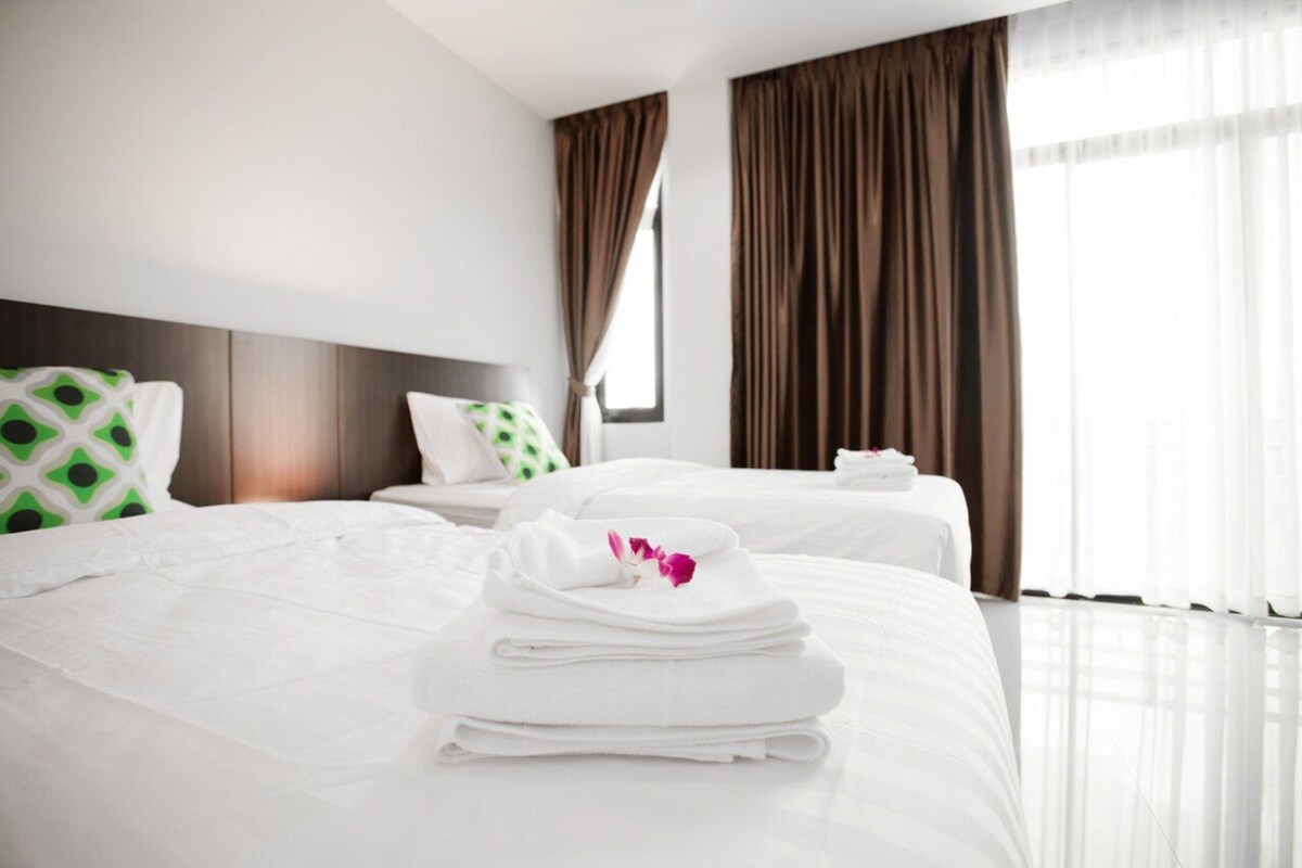 A hotel room with two beds, a large window with curtains, and a neatly made bed with towels and a flower.