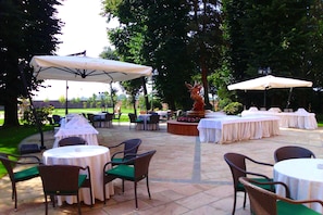 Un espace extérieur aménagé en terrasse, avec des tables et des chaises rondes, protégé par des parasols blancs, et agrémenté d’une fontaine décorative.
