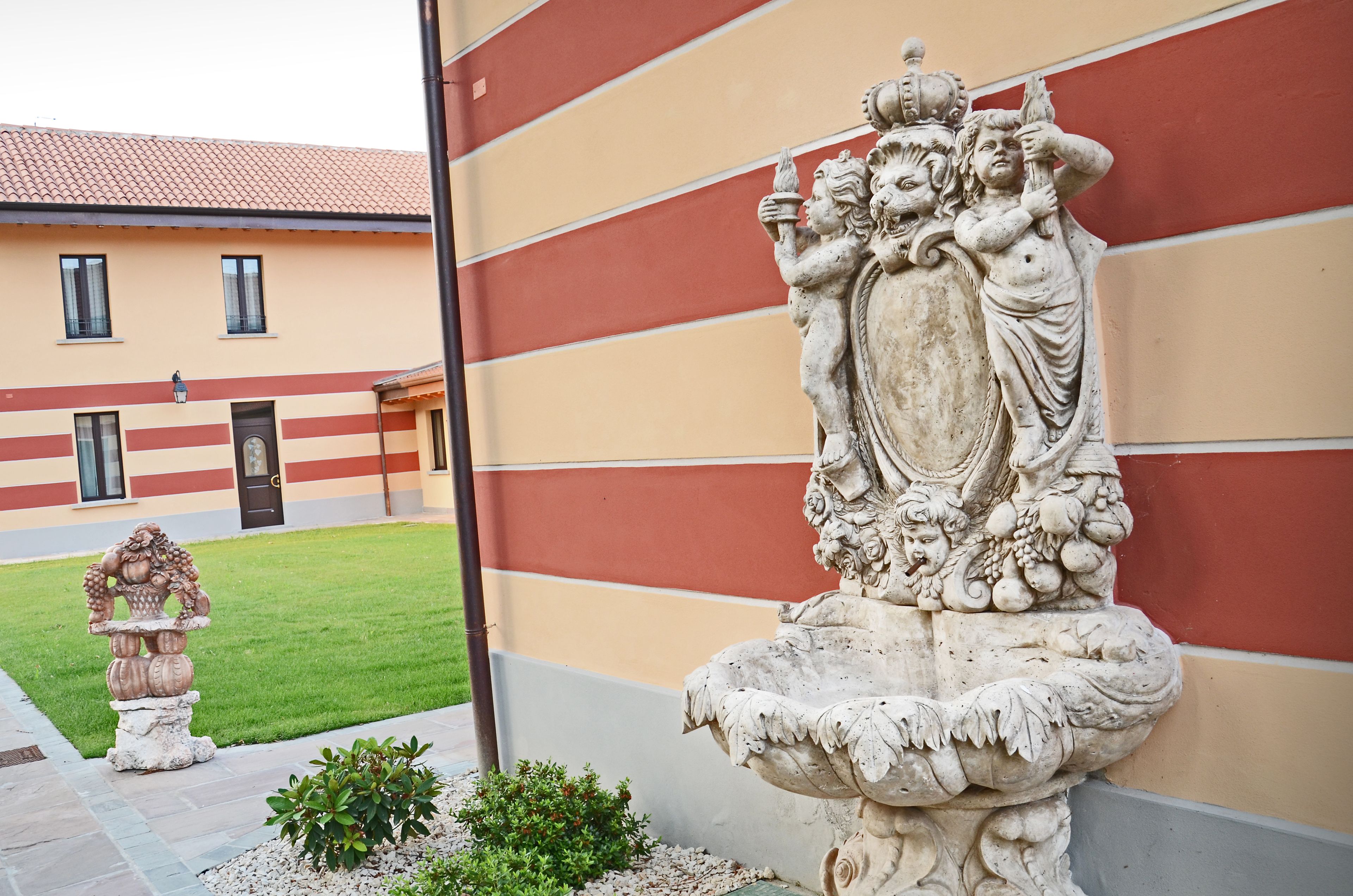 A courtyard with a stone fountain and a statue, a building with a striped facade, and a well-maintained lawn.
