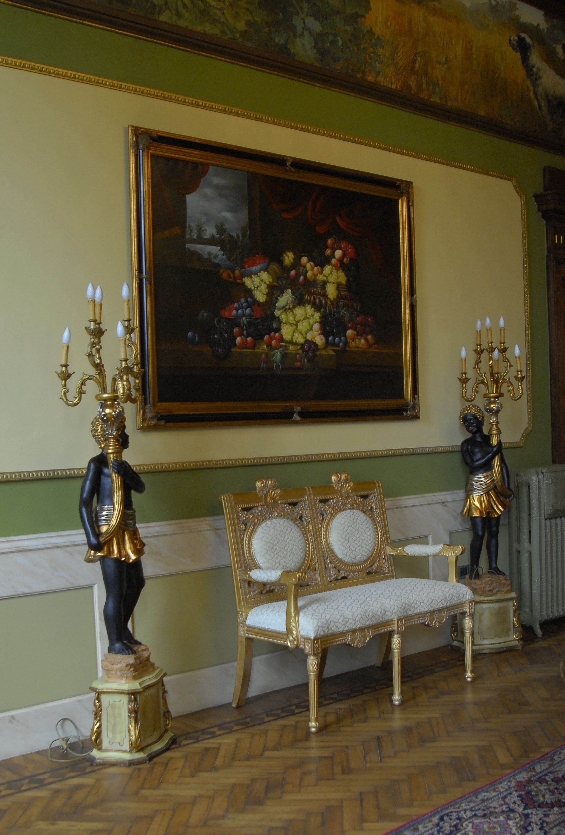 A room with a large painting of fruit, a gold-framed mirror, and two statues on pedestals.