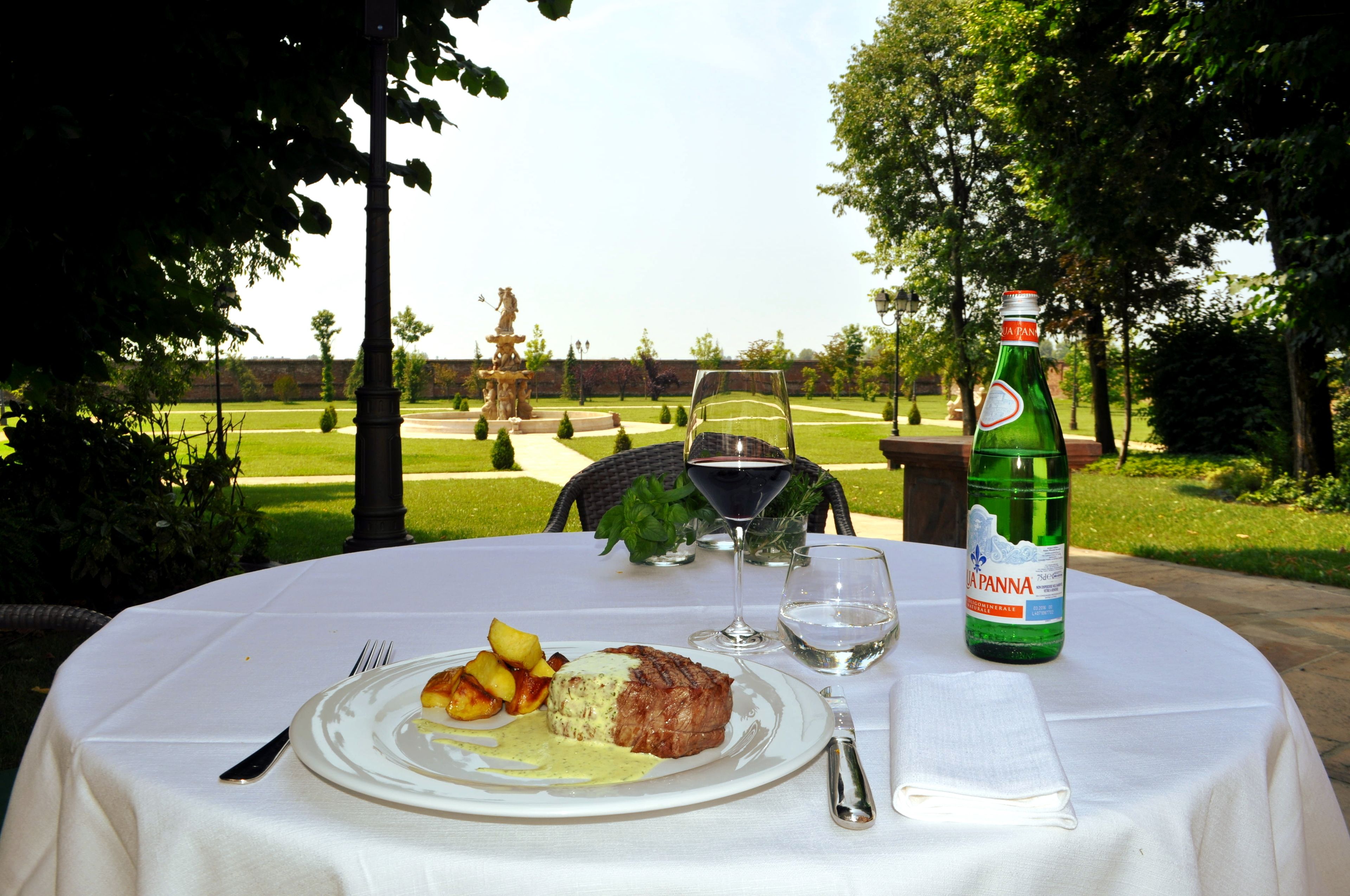 A table set outdoors with a bottle of La Panna wine, a plate with steak and potatoes, and a view of a park with a statue.