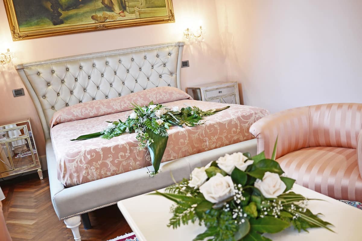 A bedroom with a tufted headboard, a bed with a pink patterned bedspread, a bedside table with a lamp, and a pink armchair.