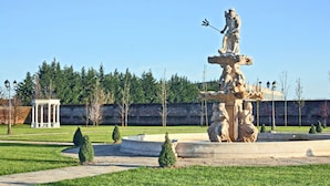 Une fontaine classique ornée de statues, entourée d’une pelouse bien entretenue et d’un mur de briques en arrière-plan.