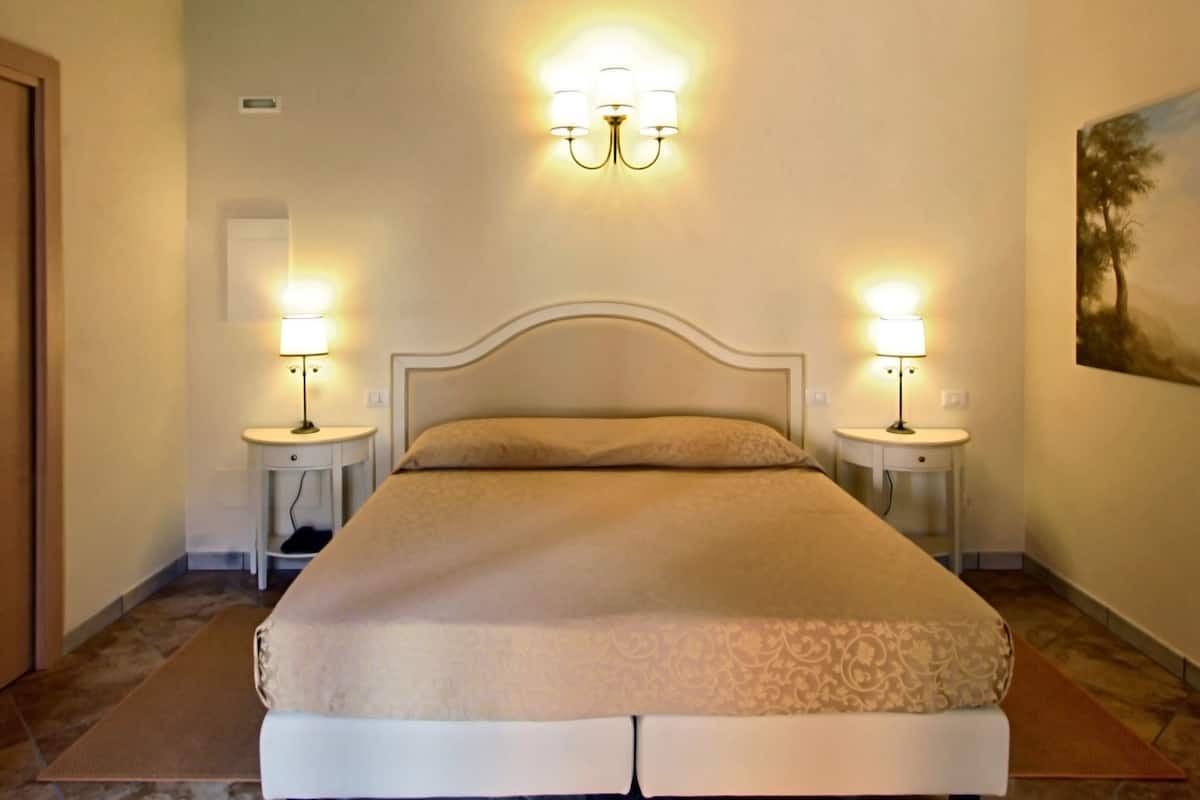 A bedroom with a large bed, wooden ceiling beams, two bedside lamps, and a painting on the wall.