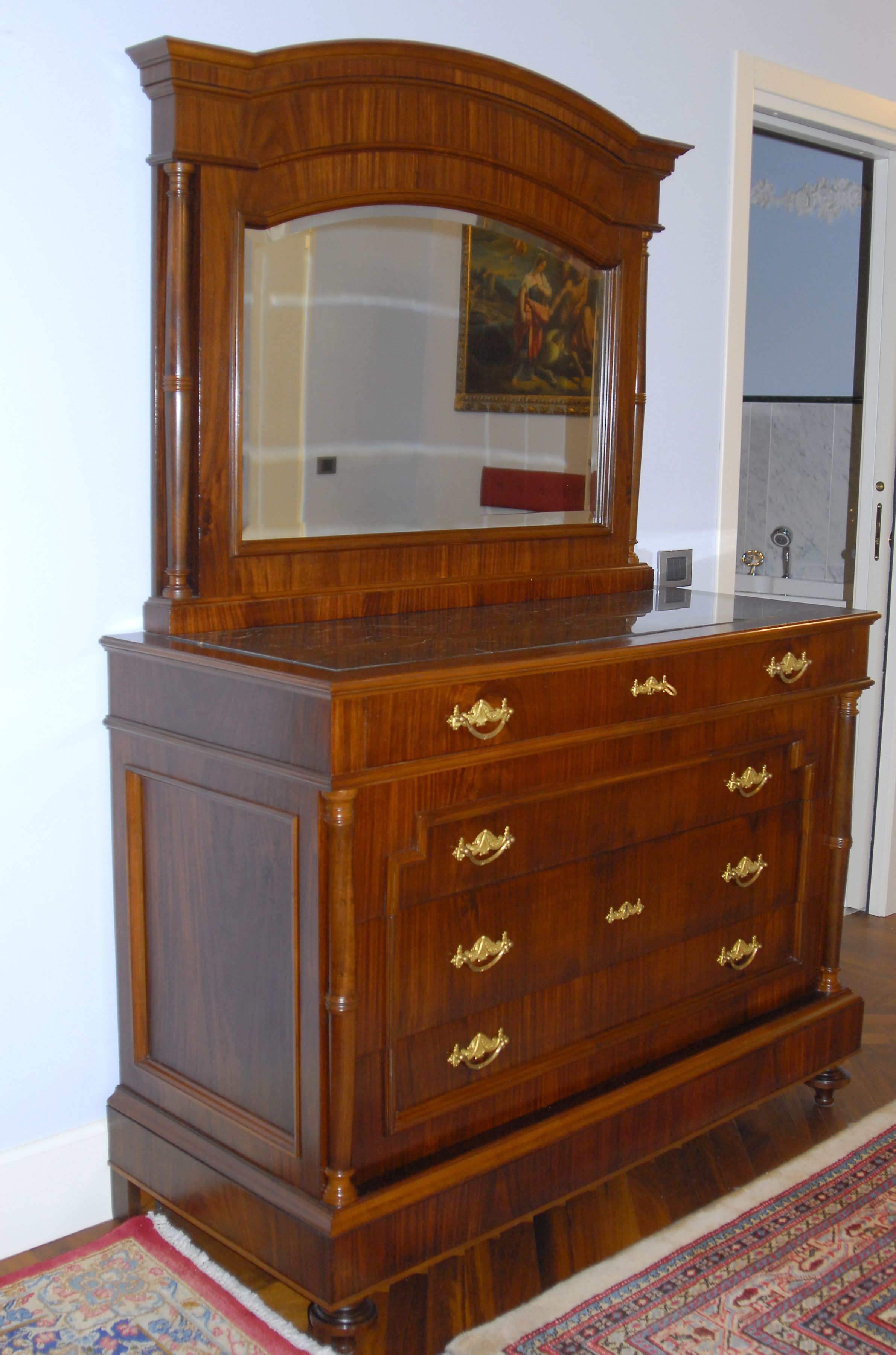 Une commode en bois avec un miroir et plusieurs tiroirs, placée dans une pièce où se trouve un tapis à motifs et une porte en arrière-plan.