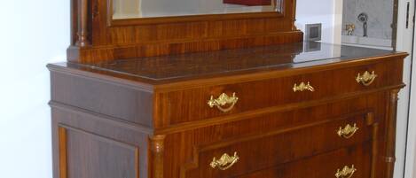 Une commode en bois avec un miroir et plusieurs tiroirs, placée dans une pièce où se trouve un tapis à motifs et une porte en arrière-plan.