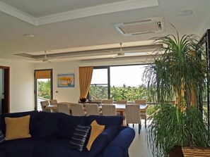 A modern living room with a dark blue sofa, a dining area with a table and chairs, and a large window offering a view of greenery.