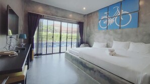 A hotel room with a large bed, a desk, a chair, a TV, and a view of a pool and greenery. - The Bike Loft Family Hostel (Hua Hin)