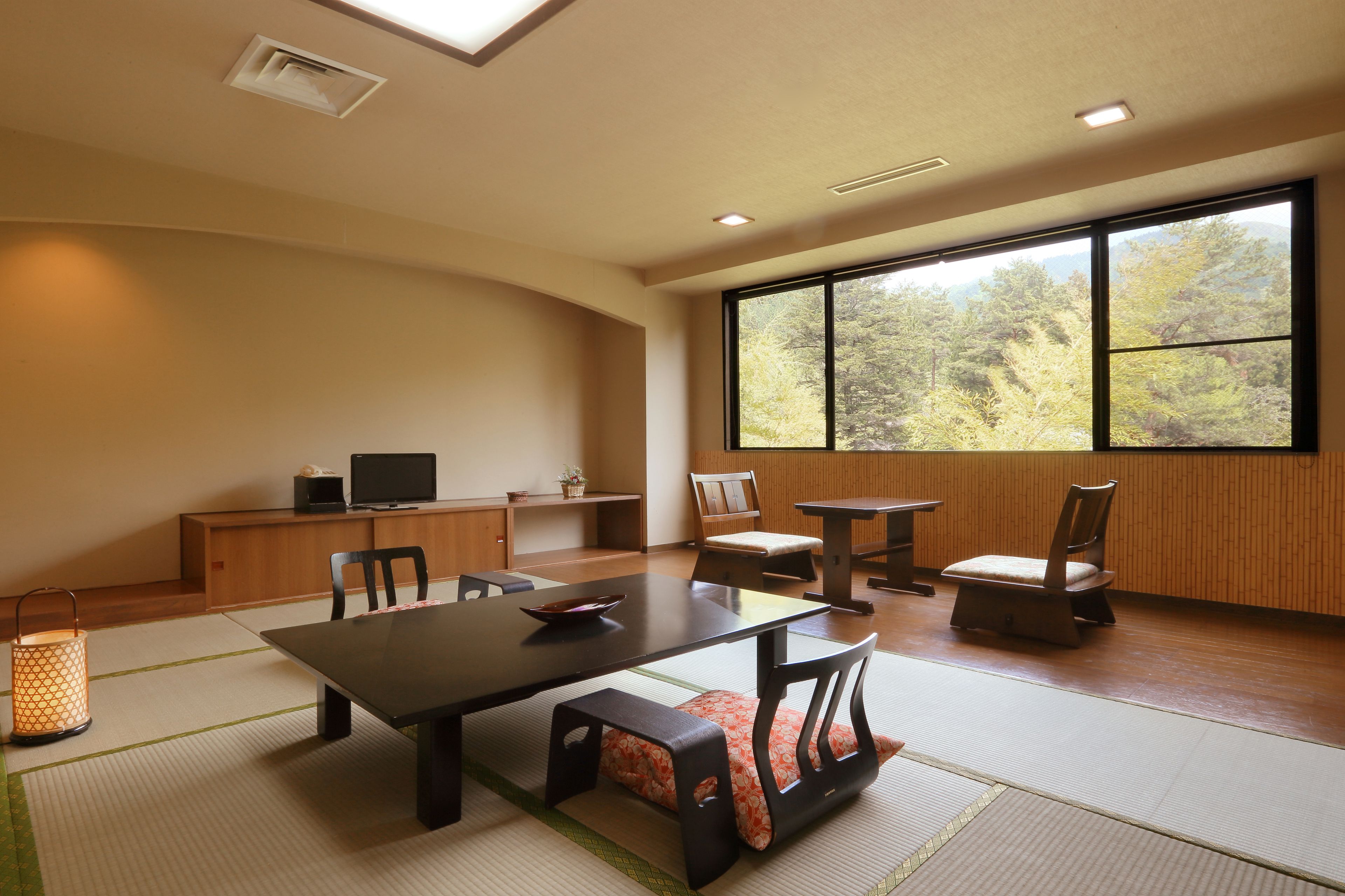 Um quarto amplo com uma grande janela, uma mesa de jantar, cadeiras, uma televisão e um tradicional candeeiro japonês.