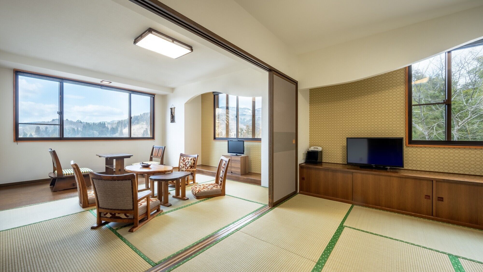 A traditional Japanese room with tatami flooring, a low table, chairs, a television, and large windows offering a view of the outdoors.