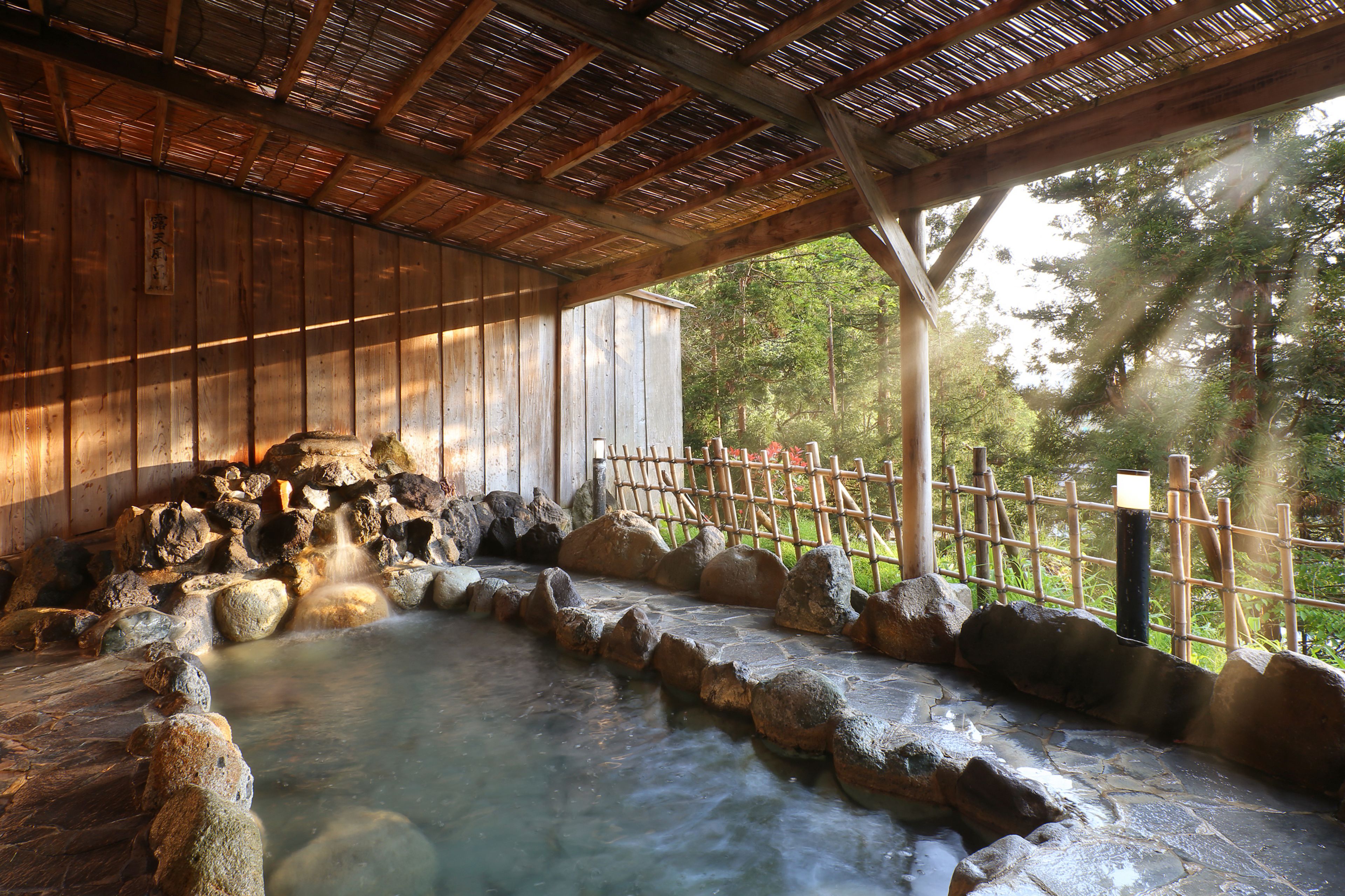 Un balneario de madera tradicional con un área de aguas termales de piedra, vigas de madera y una cerca de bambú.