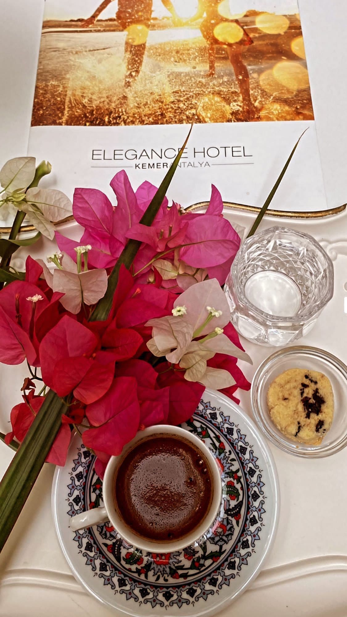 Una mesa servida con una taza de café, un vaso de agua, una galleta y un ramo de flores, junto con un menú del Hotel Elegance.