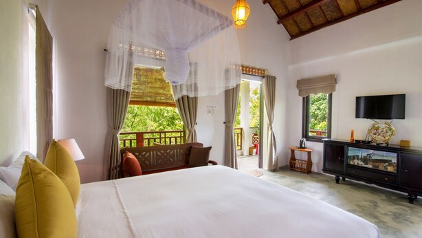 A spacious bedroom with a large bed, a television, and a balcony with a view of greenery. - Blue Beach Village Homestay (Da Nang)