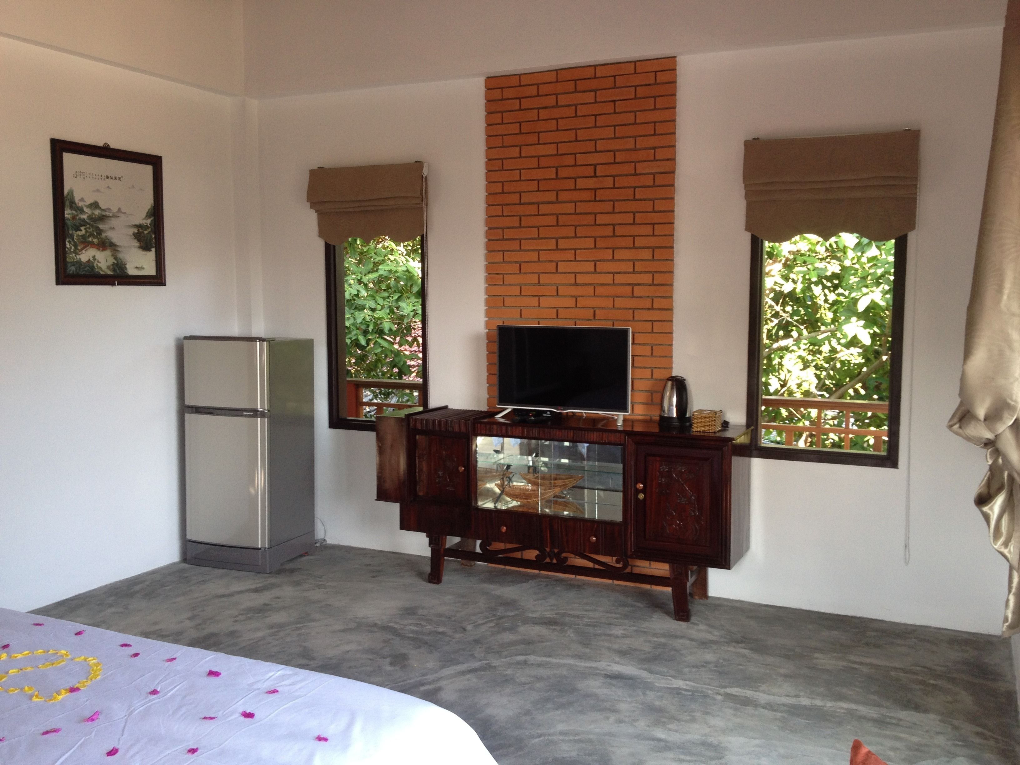 A bedroom with a bed, a TV on a wooden stand, a refrigerator, and windows with curtains.