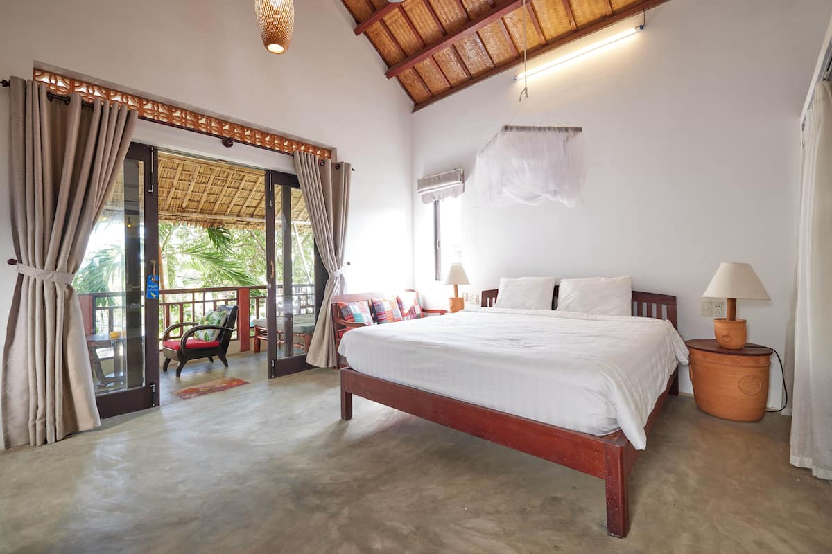 A spacious bedroom with a large bed, a wooden ceiling, and a view through a glass door to an outdoor area with a chair and a table.