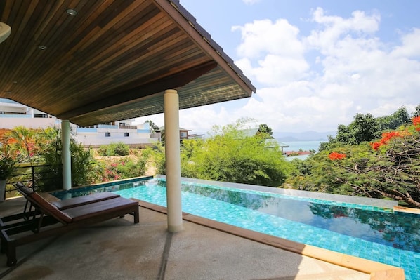 A rooftop infinity pool with a wooden pergola, surrounded by lush greenery and a view of the sea.
