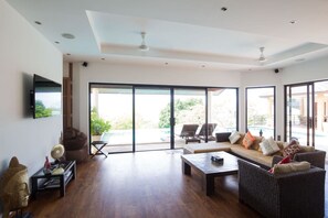 A spacious living room with a large sofa, a coffee table, and a television mounted on the wall. There are large windows providing a view of the outdoors.