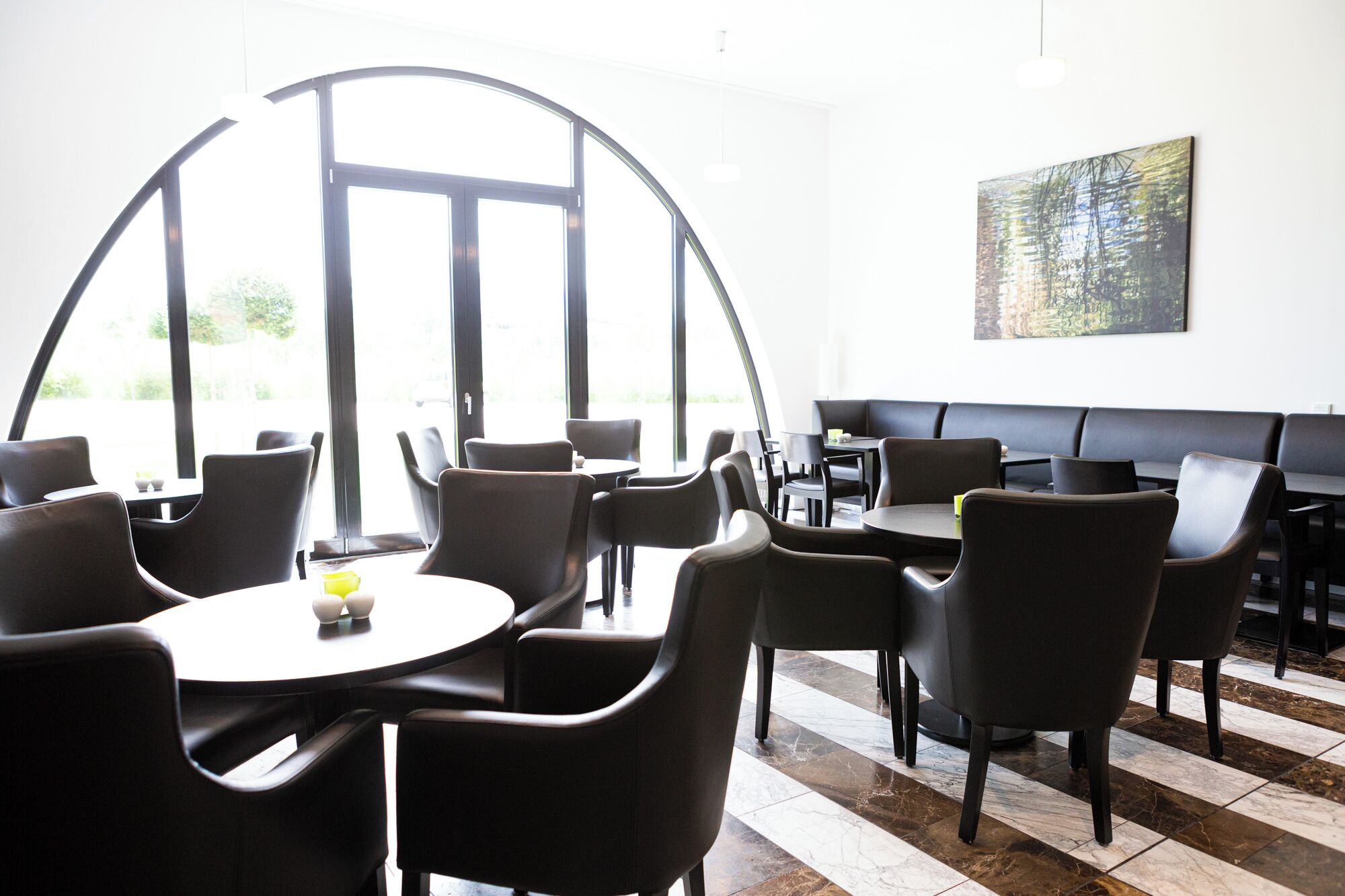 A modern restaurant with round tables and black chairs, large windows, and a painting on the wall.