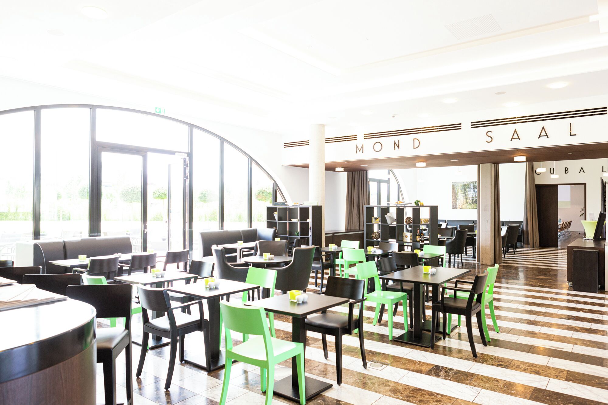 Un moderno comedor interior con mesas y sillas, ventanas amplias y un letrero que dice "MOND SAAL".
