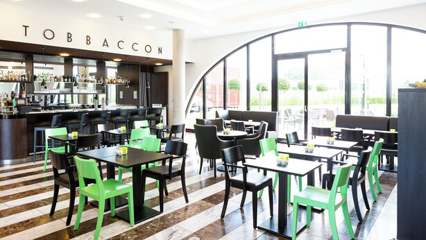 A restaurant with a bar area, seating with green and black chairs, and a view of the outside. - Hotel Tobbaccon (Bensheim)