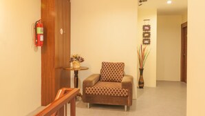 A hallway with a wooden door, a fire extinguisher, a patterned armchair, a small round table with a vase, and a wall-mounted light fixture. - M Y Home Hotel (Chiang Mai)